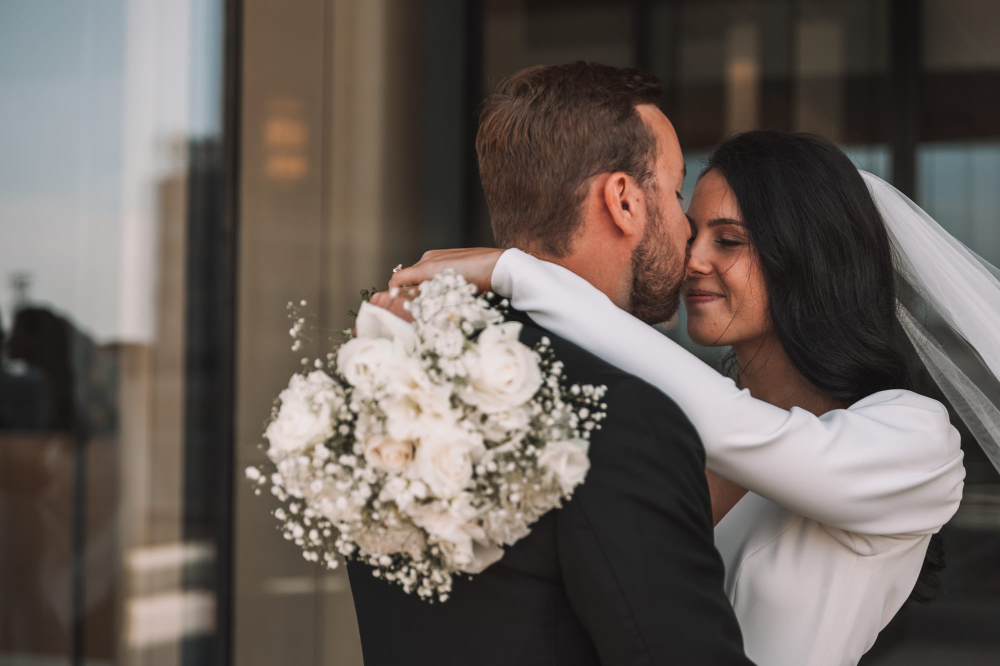 Groom kissing the bride on the forehead while holding a white bouquet